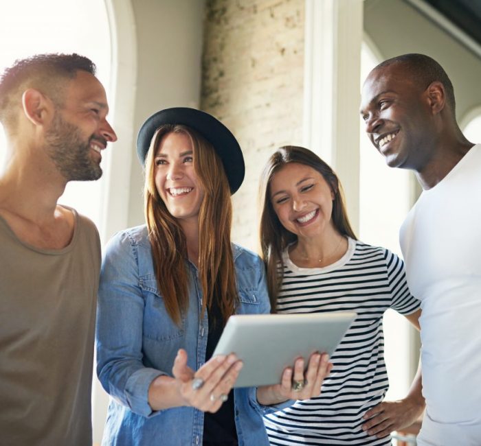 Group of four young stylish friends laughing and watching something on tablet in light modern room.