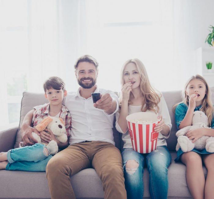 Fun time together. Happy family of four is watching interesting educational documentary, eating popcorn, at home on a couch