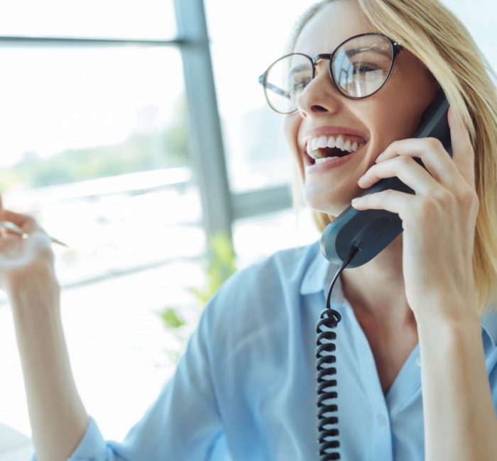 Overflowing with happiness. Overjoyed pretty woman sitting in her office and laughing happily while talking on the landline phone